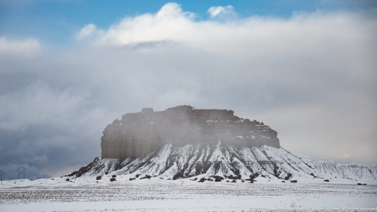 A flat-topped sandstone butte rises from a snowy plain, its slopes marked by white drainage lines beneath low clouds.