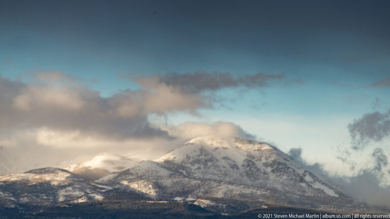 A snow-covered mountain peak rises above dark foothills and is partly wrapped in cloud.