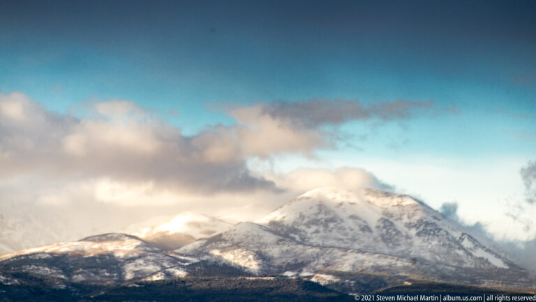 Clouds drift across a snowy summit above a series of dark ridges and foothills.