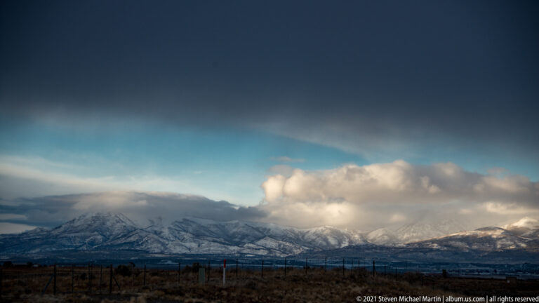 A snow-covered mountain range stretches beneath a dark blue sky, with a bright cloud over the peaks.
