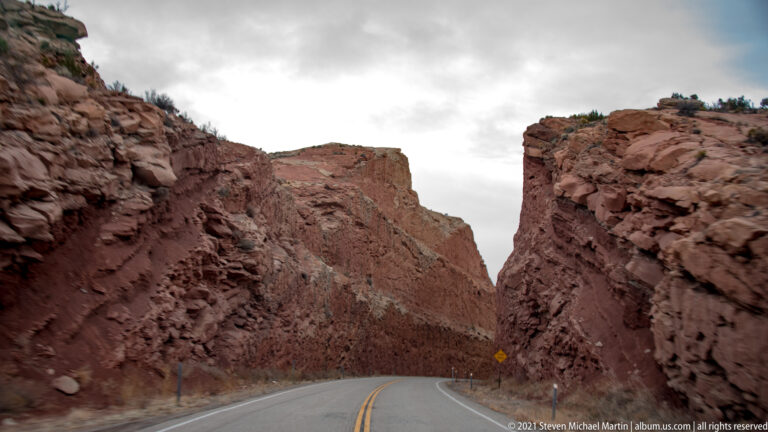 A two-lane road curves between high red sandstone walls, marked by a yellow warning sign.