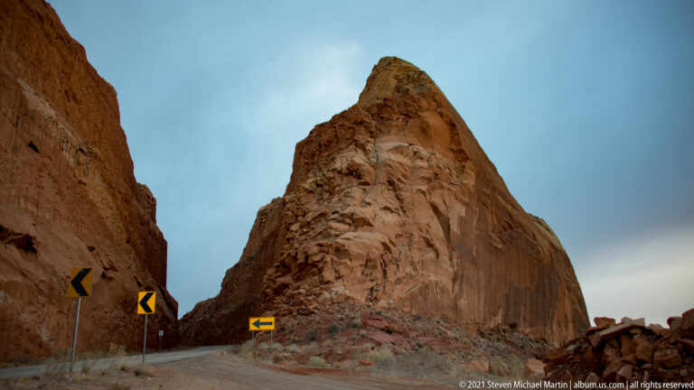 A pointed sandstone pinnacle towers beside a mountain road marked by yellow chevron signs.