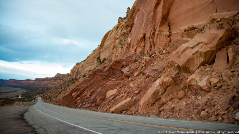 A road curves beside a fractured red sandstone wall, opening toward a broad valley.