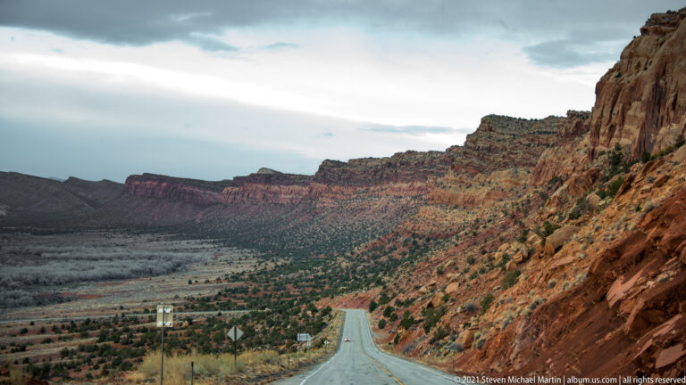 A highway descends through a valley beneath a red escarpment and distant layered mesas.