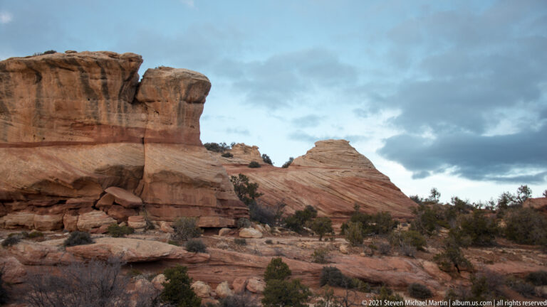 Layered sandstone outcrops rise above scattered desert shrubs beneath a cloudy sky.