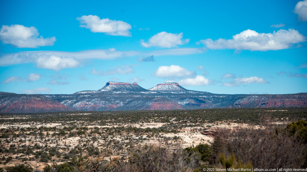 Two flat-topped Bears Ears buttes stand on the horizon beyond a snowy plateau and dark scrub.