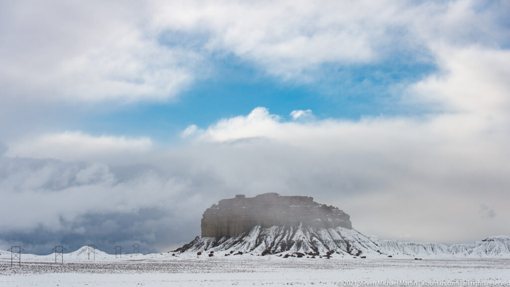 A distant mesa stands on a snowy plain beneath a broad blue opening in layered clouds.