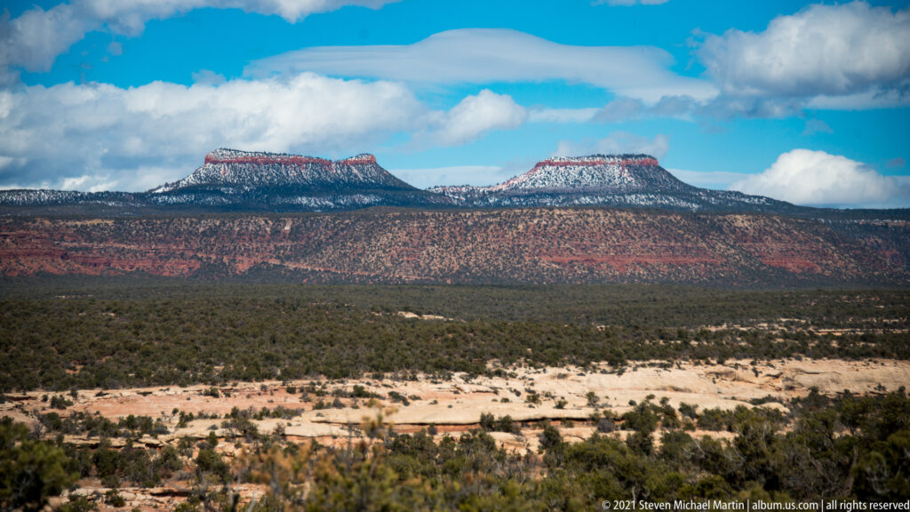 The twin Bears Ears rise above red cliffs and a broad band of dark desert woodland.