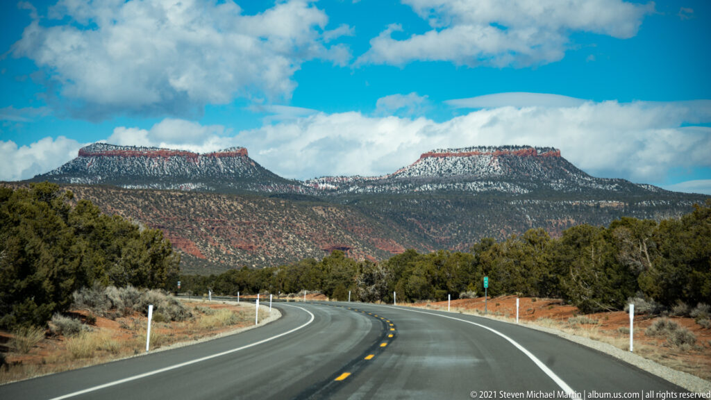 A paved road curves between juniper and scrub toward two flat-topped, red-banded mesas beneath scattered clouds.