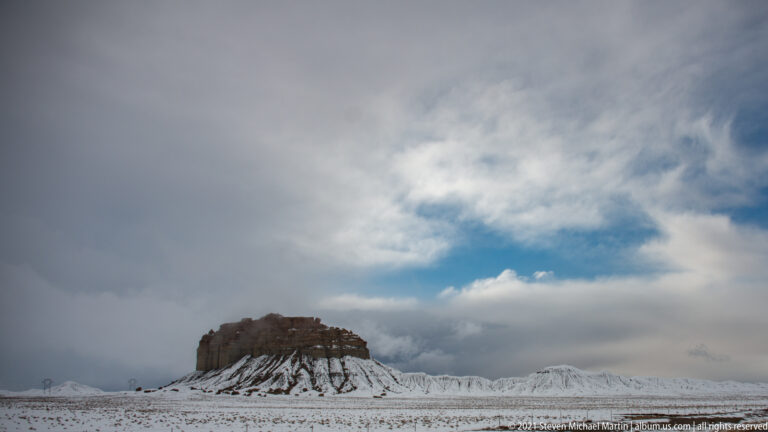 A distant butte stands left of center on a snowy plain beneath gray clouds and a patch of blue sky.