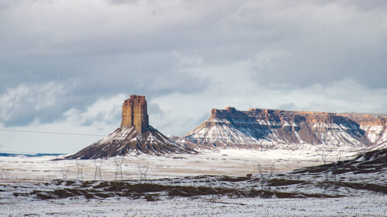 A narrow sandstone tower stands before a long mesa across a snowy basin, with utility lines crossing the foreground.