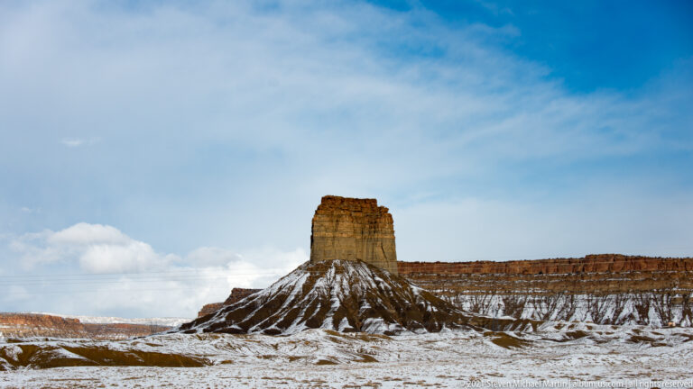 A blocky sandstone tower rises above snow-filled gullies, with a layered red escarpment behind it.