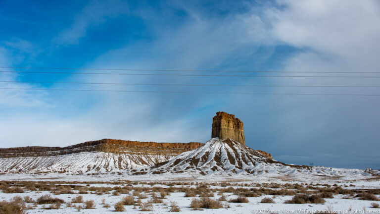 A narrow butte rises before a long mesa on a snowy plain, with utility wires crossing a blue sky.