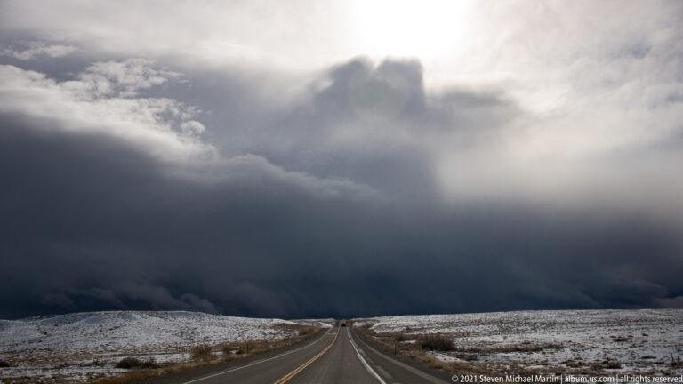 A highway runs across a snowy plateau toward a dark storm bank beneath a pale sun.