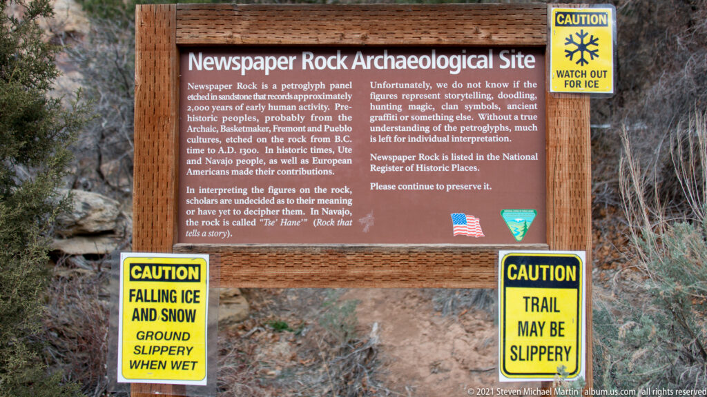 A wooden Newspaper Rock interpretive sign stands among rocks and brush, flanked by yellow winter trail warnings.