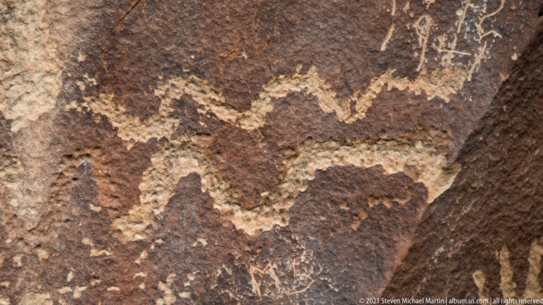 A pale winding line and smaller petroglyph marks cross a dark sandstone surface.