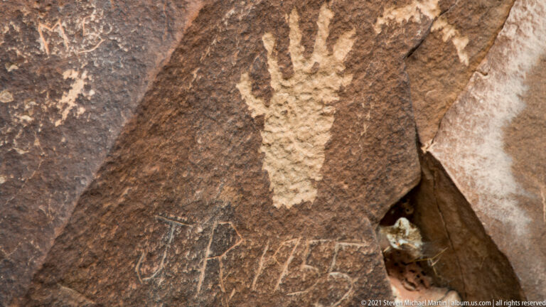 A tall pale petroglyph appears on a sloping dark panel beside a deep rock seam.