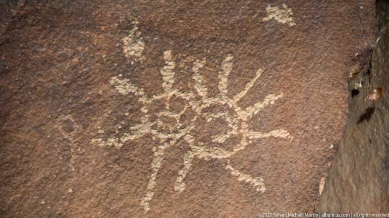A pale petroglyph with a central body and radiating lines stands alone on dark sandstone.