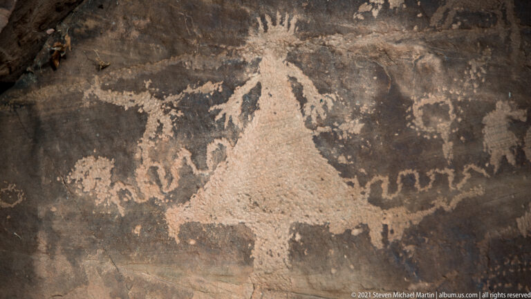 A large pale radiating figure rises above smaller petroglyph forms and a wavy line.