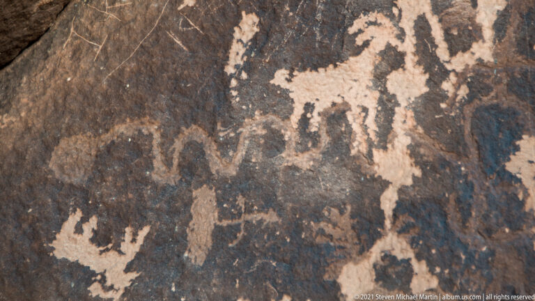 Pale petroglyph fragments and small upright forms appear across a dark mottled rock face.