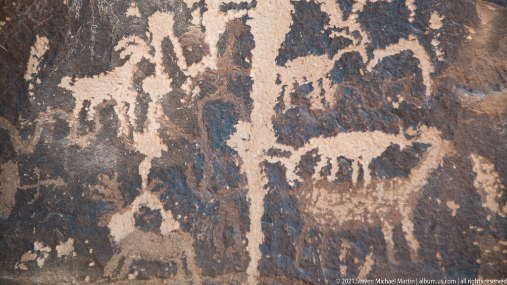 Large pale figures and smaller linear petroglyphs cover a patchy dark sandstone panel.