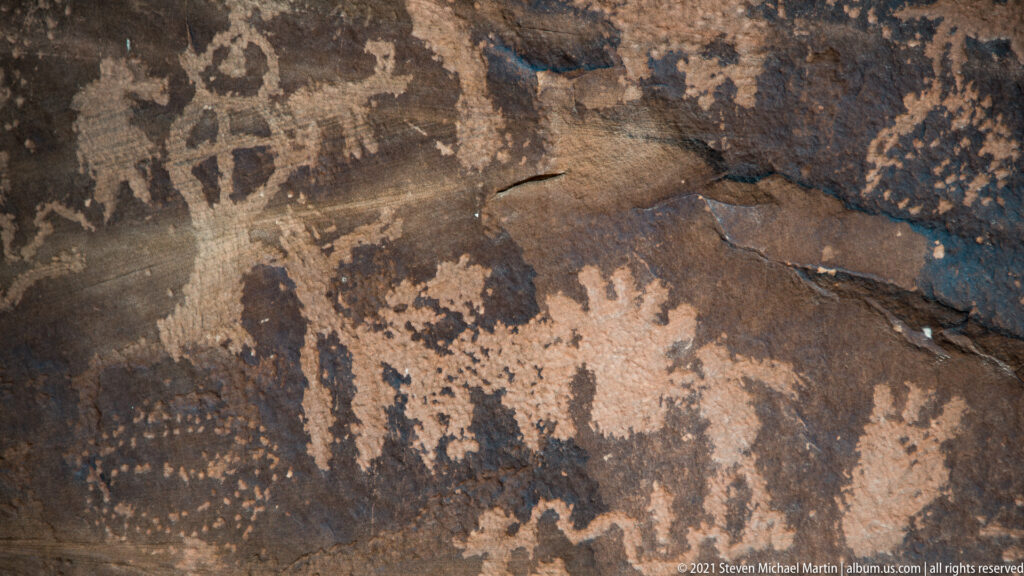 A broad pale four-legged form is surrounded by smaller petroglyphs on cracked sandstone.