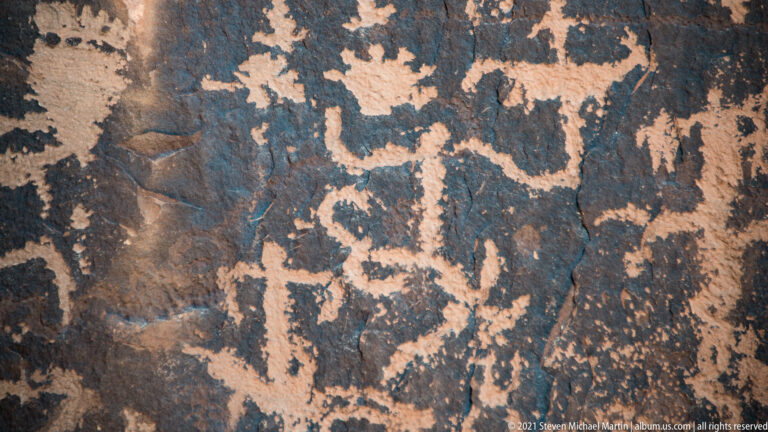 Many pale figure-like and curving petroglyph marks cover a dark sandstone surface.