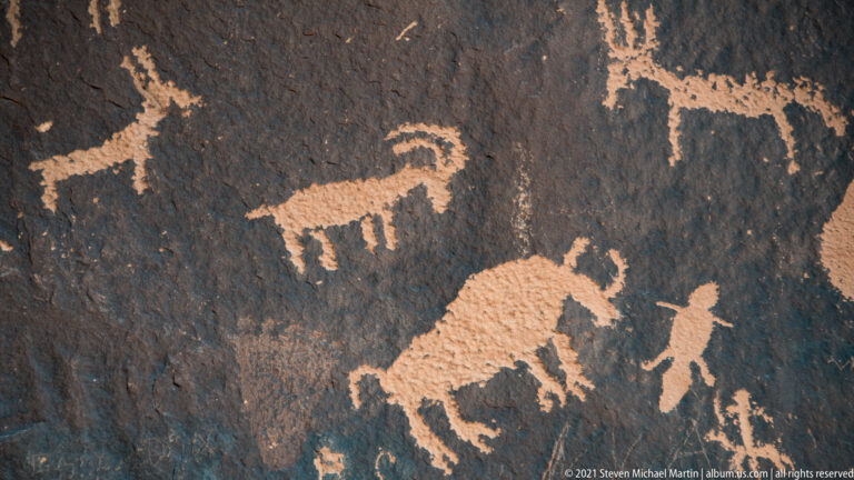 A large quadruped-like petroglyph sits below smaller upright figures on dark rock.