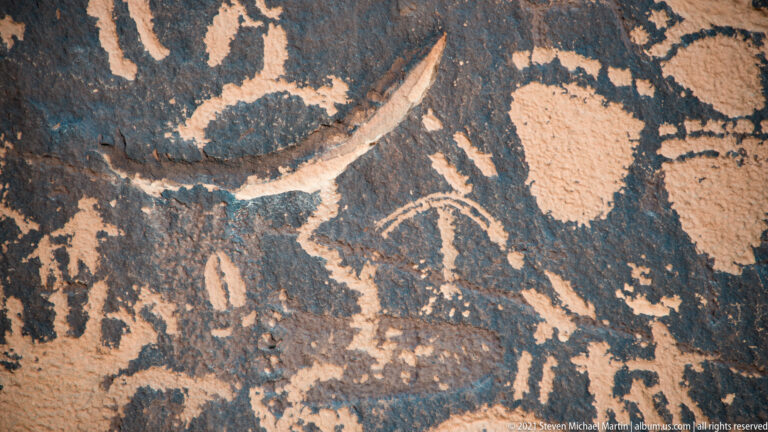 A sweeping pale curve crosses above a broad figure and smaller marks on dark sandstone.