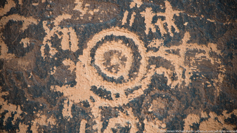 A pale concentric circle with a central mark is surrounded by smaller petroglyph lines.