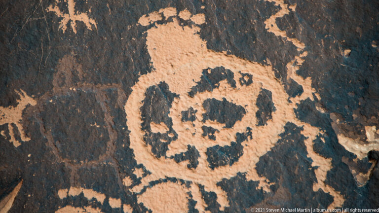 A large ringed, face-like petroglyph fills the center of a dark rock panel, with smaller figures around it.