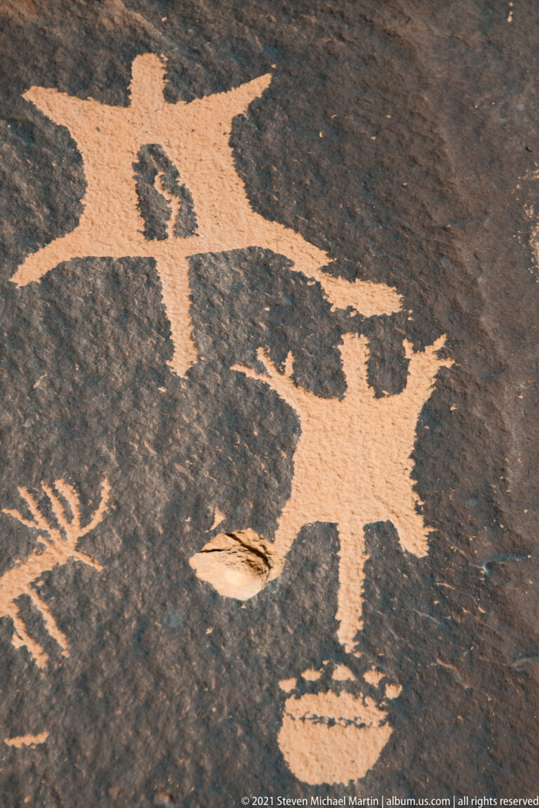 A broad-limbed human-like figure spans a dark stone panel, with smaller petroglyph fragments along the left edge.