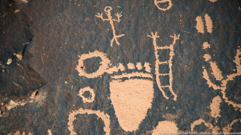 A ladder-like petroglyph stands beside a large rectangular-bodied form and smaller circles on dark stone.