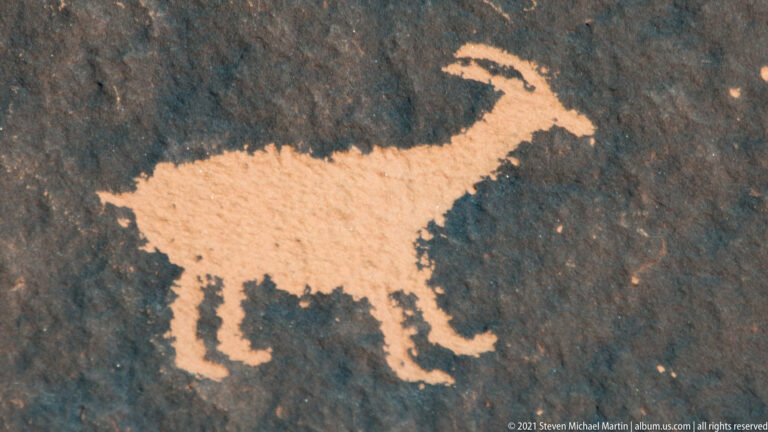 A pale quadruped with an elongated body and raised, curved head is carved in profile on dark stone.