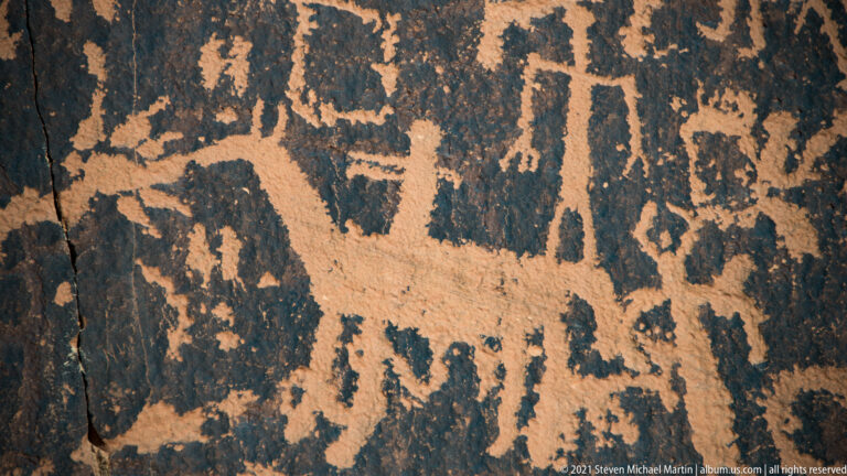 A large quadruped lies among upright figures and abstract marks on a densely carved rock surface.