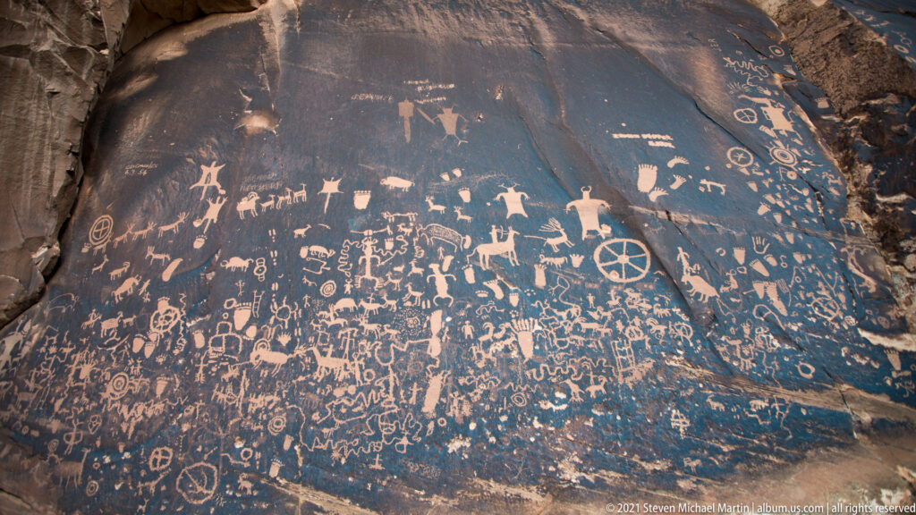 A dense petroglyph grouping includes small figures and a large divided circle on sloping dark sandstone.