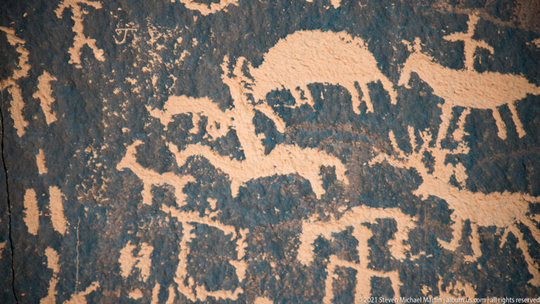 A broad quadruped with curved head marks spans above a dense band of smaller petroglyphs.