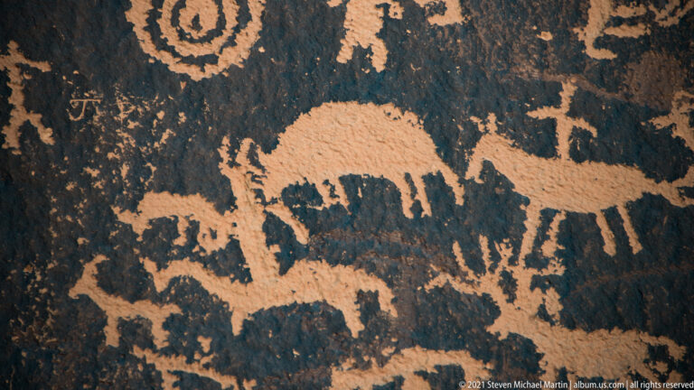 A long-bodied quadruped sits within a crowded field of smaller animal-like and human-like petroglyphs.