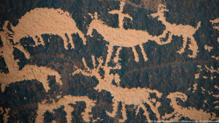 A large quadruped with a raised head appears above smaller petroglyph figures on mottled dark stone.