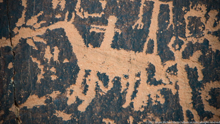 An elongated pale petroglyph spans above a narrow upright figure and smaller shapes on dark rock.