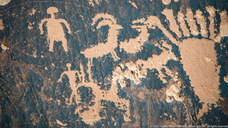 A rectangular-bodied figure, smaller upright forms, and an animal-like motif share a weathered rock panel.