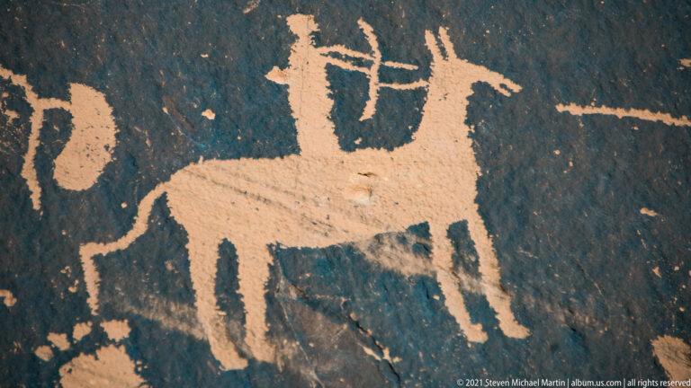 A large quadruped with branching head marks stands above smaller petroglyphs on dark rock.