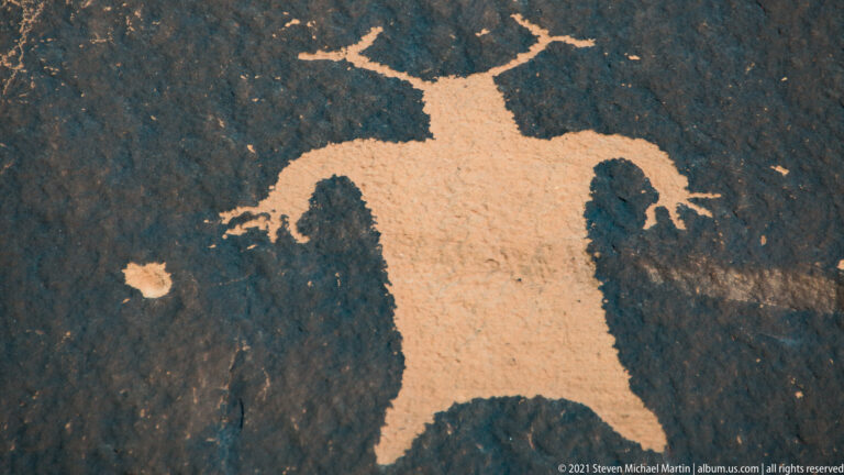 A broad-bodied figure extends long arms across a dark rock surface, with legs below.
