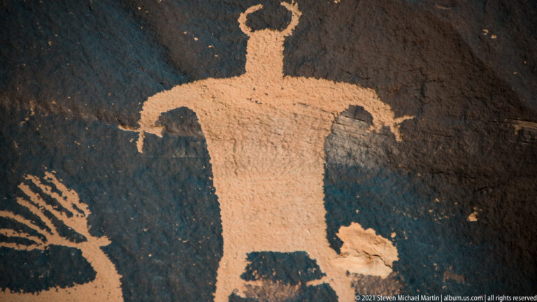 A front-facing figure with wide arms and a loop above its head is carved into dark stone.