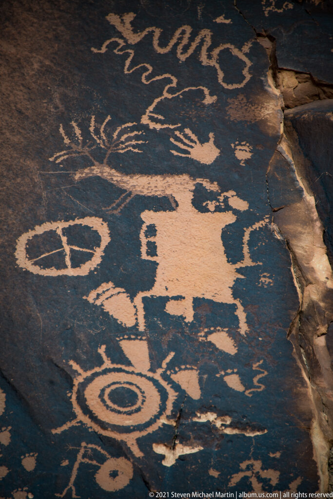 A large central petroglyph sits among circles and elongated figures on a broad slab beside a fractured edge.
