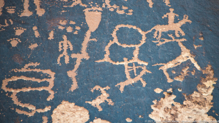 Large looped petroglyphs with short crossbars sit beside upright figures and smaller circles on dark rock.