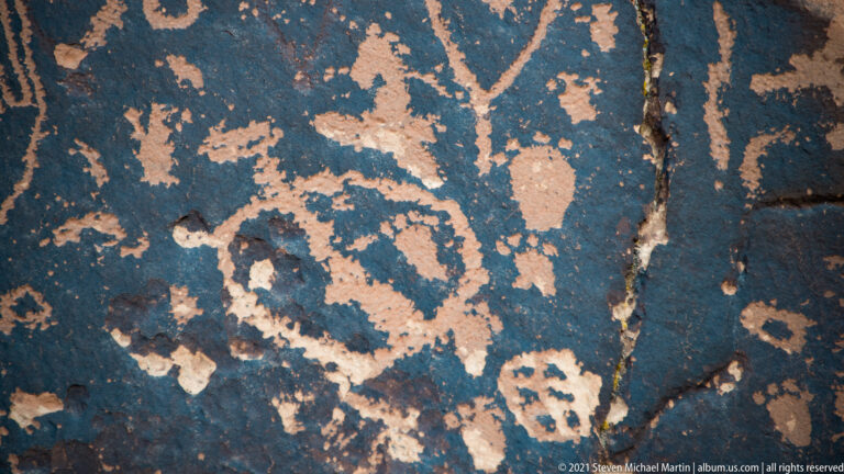 Overlapping pale petroglyphs cover dark sandstone, including figure-like outlines, dots, and branching forms.