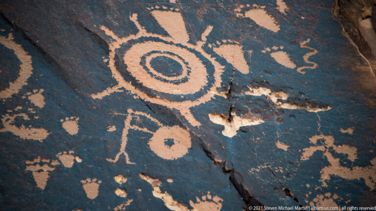A large concentric circle and smaller figure-like carvings appear on dark sandstone beside a natural crack.