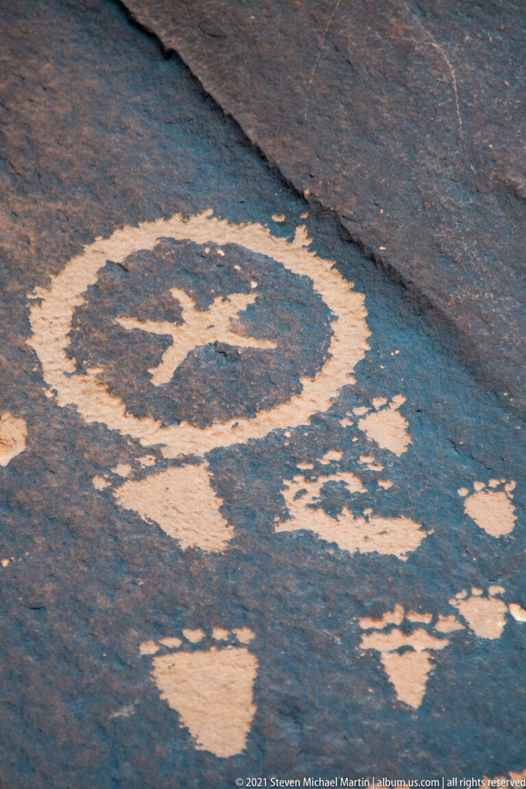 A broad pale ring-shaped petroglyph stands out on a sloping dark sandstone surface with scattered marks and a crack.