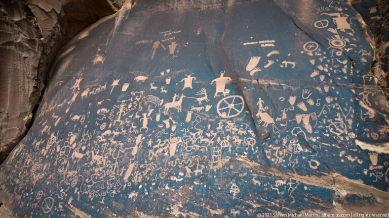 A curved sandstone panel carries many petroglyphs, including human-like figures, animals, circles, and a wheel-like form.